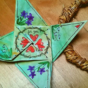 Può includere: Una stella di origami di carta verde con una ghirlanda di viti e cuori rossi al centro. La stella è decorata con fiori viola e bianchi e le parole "for you".