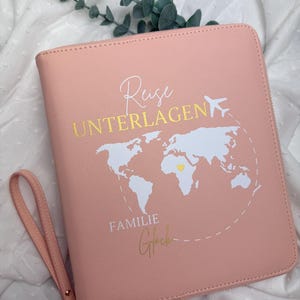 Könnte beinhalten: Ein rosafarbenes Reise-Dokumenten-Etui mit goldenem Reißverschluss und Handschlaufe. Auf der Vorderseite befindet sich eine weiße Weltkarte mit goldenem Herz und den Worten "Reise UNTERLAGEN" und "FAMILIE Glück" in goldener Schrift.