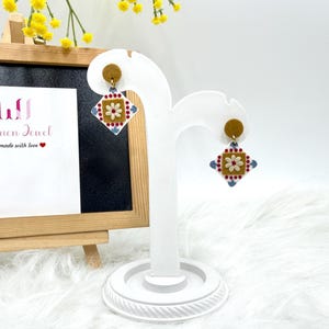 May include: A pair of earrings with a geometric design. Each earring features a small, round stud in a tan color, with a square pendant below. The pendant has a tan center with a white flower and red and blue accents. The earrings are displayed on a white stand.