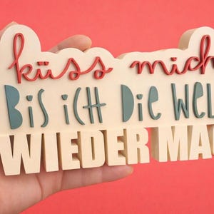 Op de afbeelding: Een crèmekleurig houten bord met de Duitse zin "Küss mich, Bis ich Die Welt Wieder Mag" in rode en blauwgroene letters. Het bord wordt tegen een rode achtergrond gehouden.