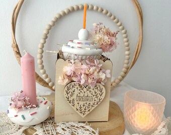 May include: A decorative birthday-themed arrangement featuring a cupcake-shaped centerpiece with a lit orange candle, surrounded by pink flowers. A wooden heart with text is attached to a wooden box. A pink candle and a glass candle holder are also present.