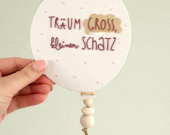 May include: A white, oval-shaped decorative piece with the German text "TRÄUM GROSS, kleiner SCHATZ" in brown lettering. The design includes a cloud graphic and small dotted accents. A string of wooden beads and a tan cord hang from the bottom.