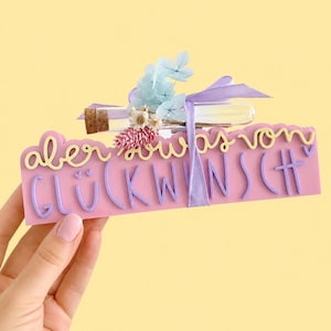 May include: A pink rectangular sign featuring the yellow and purple text "aber sowas von GLÜCKWUNSCH" with two small hearts. A clear glass vial with a cork stopper is tied to the top with a light purple ribbon, decorated with dried blue and pink flowers.