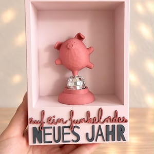 May include: A pink rectangular shadow box containing a small pink pig figurine balanced on a silver mirrored disco ball. The base of the box features the text "auf ein funkelndes NEUES JAHR" in a combination of cursive and block lettering.