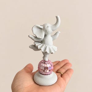 Op de afbeelding: Een lichtgrijs olifantbeeldje met een geplooid tutu, balancerend op een roze gespiegelde discobal. De olifant heeft de slurf omhoog en de oren wijd gespreid, staand op een ronde voet die de reflecterende roze bol ondersteunt.