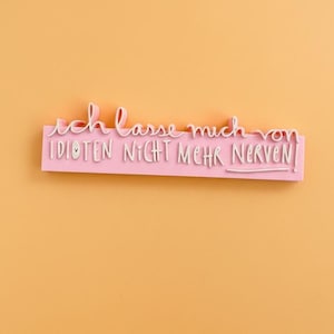 Könnte beinhalten: Ein rosa rechteckiges Schild mit dem deutschen Satz "Ich lasse mich von Idioten nicht mehr nerven!" in weißer Schrift. Das Schild befindet sich vor einem orangefarbenen Hintergrund.