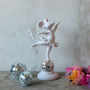 Könnte beinhalten: Eine weiße Mausfigur in Ballettpose mit Tutu, die auf einer kleinen Spiegelkugel balanciert. Die Maus steht auf einem runden weißen Sockel. Zwei weitere Spiegelkugeln und ein Bündel bunter Trockenblumen sind ebenfalls im Bild zu sehen.