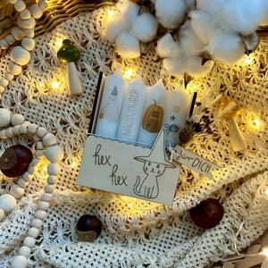 May include: A decorative wooden box with the words "hex hex" and a cat wearing a witch hat. The box contains three candles with illustrations and the word "NACHTZAUBER". The scene includes decorative elements like cotton, beads, and chestnuts.