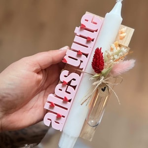 Könnte beinhalten: Eine dekorative Kerze mit dem Namen "Alessia" in rosa Buchstaben, akzentuiert mit kleinen roten Herzen. Die Kerze ist weiß und mit Trockenblumen und einem kleinen Glasröhrchen verziert. Die Kerze ist an einer Holzbasis befestigt.