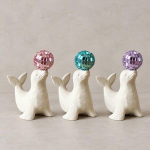 Könnte beinhalten: Drei cremeweiße Robbenfiguren, die jeweils eine kleine, runde, reflektierende Discokugel auf der Nase balancieren. Die Discokugeln sind in den Farben Pink, Türkis und Violett gehalten. Jede Robbe sitzt in einer aufrechten Position mit erhobenem Schwanz.