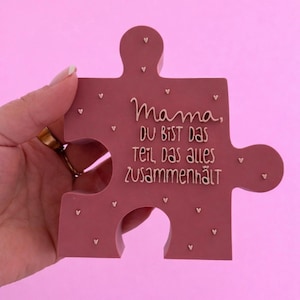 Puede incluir: Una pieza de rompecabezas malva con el texto alemán "Mama, du bist das Teil, das alles zusammenhält" en letras color crema. Pequeños corazones blancos están dispersos por la superficie. La pieza del rompecabezas se sostiene sobre un fondo rosa.