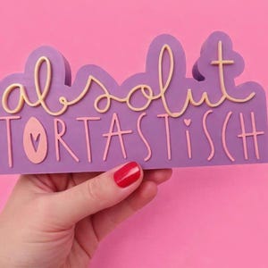 Op de afbeelding: Een paars bord met de woorden "absolut TORTASTISCH" in een mix van script- en blokletters. Het bord heeft een wolkvorm en wordt tegen een roze achtergrond gehouden. De letters zijn lichtbeige.