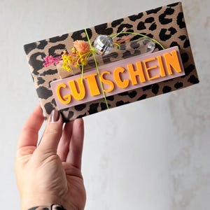 Op de afbeelding: Een rechthoekige cadeaubon verpakt in bruin papier met een zwart luipaardpatroon. De voorzijde is voorzien van een lichtroze rechthoekig plaatje met de oranje tekst GUTSCHEIN, versierd met een kleine zilveren discobal en gedroogde bloemen.
