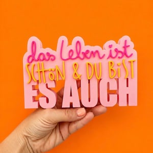 May include: A pink sign with raised lettering featuring the German text das leben ist SCHÖN & DU BIST ES AUCH. The top line is in a pink cursive script, the middle line is in yellow block letters, and the bottom line is in large pink block letters.