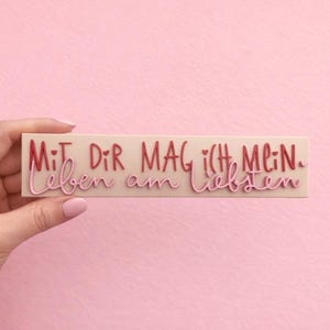 Puede incluir: Un objeto rectangular blanquecino con la frase alemana "MIT DIR MAG ICH MEIN Leben am liebsten" en escritura roja y rosa. El objeto se sostiene sobre un fondo rosa.