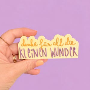 Puede incluir: Una pegatina amarilla clara con la frase alemana "danke für all die kleinen Wunder" en escritura naranja y morada. La pegatina se sostiene sobre un fondo morado.