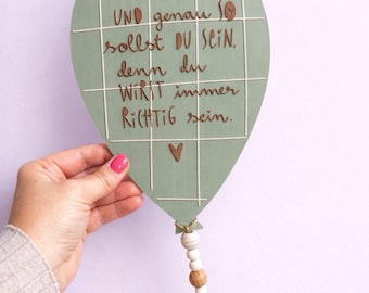 May include: A sage green balloon-shaped wooden plaque featuring a white grid pattern and brown text that reads: DU BIST DU, UND genau SO sollst DU SEiN, denn du WIRST immer RiCHTiG sein. A small brown heart and a beaded jute string dangle below.