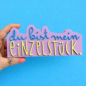 Könnte beinhalten: Ein lavendelfarbenes Schild mit dem deutschen Satz "Du bist mein Einzelstück" in blauen und gelben Buchstaben. Das Schild wird vor einem blauen Hintergrund gehalten. Der Satz bedeutet "Du bist mein Einzelstück."