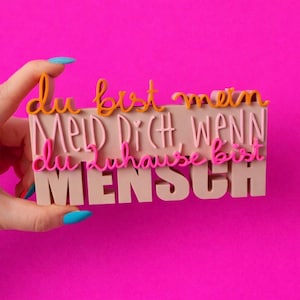 Peut inclure: Un bloc rectangulaire beige avec le texte allemand "Du bist mein Mensch, meld dich wenn du zuhause bist" en lettres orange et rose. Le bloc est tenu sur un fond rose vif.