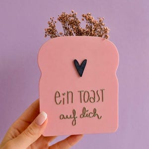 Könnte beinhalten: Ein rosa, toastförmiges Dekorationsobjekt mit einem schwarzen Herz und dem deutschen Text "ein toast auf dich". Das Objekt enthält Trockenblumen. Der Hintergrund ist einfarbig lila.