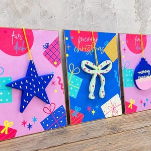 Op de afbeelding: Drie rechthoekige wenskaarten met kleurrijke patronen van geschenkdozen en hangende ornamenten. De linkerkaart heeft een blauwe ster, de middelste kaart een lichtgroene strik en de rechterkaart een blauwe en roze kerstbal met Merry Christmas.