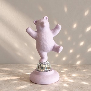 Puede incluir: Una figura de oso de color lavanda claro de pie sobre una pierna encima de una pequeña bola de discoteca plateada esférica. El oso tiene una superficie texturizada y está colocado sobre una base circular lavanda con una pose equilibrada.