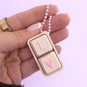 Puede incluir: Un collar con colgante beige con dos teclas de teclado cuadradas. La tecla superior tiene una "L" rosa y la tecla inferior un corazón rosa. El colgante cuelga de una cadena de cuentas rosas.