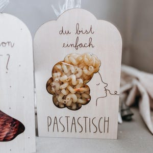 Op de afbeelding: Houten pastaverpakking met een helder venster in de vorm van een vrouwenhoofd en haar, gevuld met elleboogmacaroni. De verpakking heeft de woorden "du bist einfach" en "PASTASTISCH" in het hout geëtst.