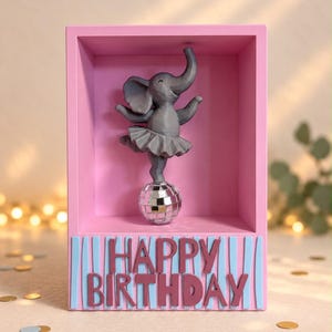 Pode incluir: Uma moldura retangular rosa contendo uma estatueta de elefante cinza vestindo um tutu e equilibrando-se sobre uma bola de discoteca prateada. A frente da moldura apresenta o texto HAPPY BIRTHDAY em letras castanhas sobre listras verticais azuis.