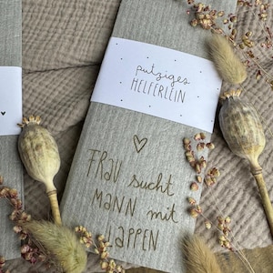 May include: A grey felt item with the text "putziges HELFERLEIN" on a white label. Below, the felt has the text "FRAU sucht MANN mit LAPPEN" with a heart symbol. Dried flowers and seed pods surround the item.