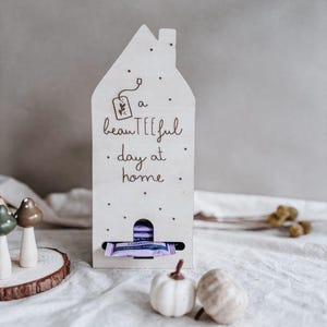 May include: A wooden house-shaped tea bag holder with the text "a beauTEEFul day at home". The holder has a slot for tea bags and is decorated with small dots and a tea bag illustration. Two small decorative pumpkins are in front of the holder.