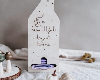 May include: A wooden house-shaped tea bag holder with the text "a beauTEEFul day at home". The holder has a slot for tea bags and is decorated with small dots and a tea bag illustration. Two small decorative pumpkins are in front of the holder.