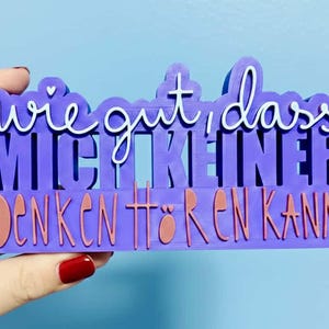 Könnte beinhalten: Ein lila Schild mit dem deutschen Text "wie gut, dass mich keiner denken hören kann" in weißer und orangefarbener Schrift. Das Schild wird von einer Hand mit rotem Nagellack vor blauem Hintergrund gehalten.