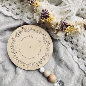Puede incluir: Un círculo de madera con un diseño floral y el texto "Die das Leben die kleinen Dinge im Leben machen" colgando de una cuerda con una cadena de cuentas. El círculo está sobre un fondo de tela gris claro.