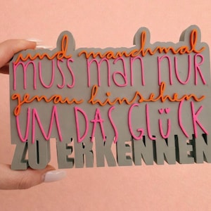 May include: A gray rectangular sign with the German text "und manchmal muss man nur genau hinsehen um das Glück zu erkennen" in orange and pink lettering. The sign is held by a hand against a pink background.