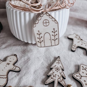 Könnte beinhalten: Weihnachtsschmuck aus Holz in verschiedenen Formen, darunter ein Lebkuchenhaus, ein Weihnachtsbaum und ein Pferd. Die Ornamente sind hellbraun mit dunkelbraunen, geätzten Details. Im Hintergrund ein weißer Keramiktopf mit Trockenblumen.