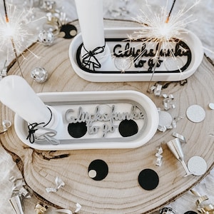 Puede incluir: Dos candelabros blancos con el texto "Glücksfunke to go" se exhiben sobre una superficie de madera, rodeados de confeti, bolas de discoteca y bengalas. Los candelabros tienen un borde negro y un lazo negro.