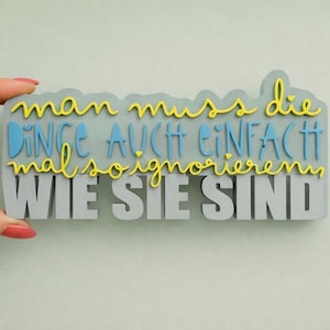 Puede incluir: Un letrero rectangular gris con la frase alemana "man muss die Dinge auch einfach mal ignorieren wie sie sind" en letras amarillas, azules y grises. El letrero está sostenido por una mano con esmalte de uñas rojo.