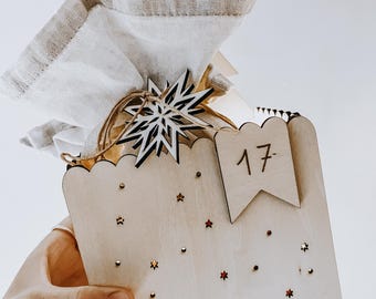 Può includere: Un calendario dell'Avvento in legno chiaro, decorato con ritagli a forma di stella e piccole stelle rosse. Un sacchetto di stoffa bianca è legato con spago e sormontato da un fiocco di neve di legno. Il numero "17" è visualizzato su una piccola bandiera.
