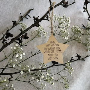 May include: A wooden star-shaped ornament with the German text "UND WIR WISSEN, DU WARST HIER, WENN DER HIMMEL NICHT SO WEIT WAR" hangs from a twine string. The ornament is surrounded by delicate white flowers and dark branches.