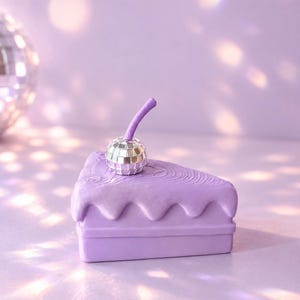 Può includere: Un portagioie a forma di fetta di torta color lavanda con una parte superiore scolpita a glassa e una ciliegia a sfera stroboscopica argentata con un gambo viola. L'oggetto ha una finitura opaca e una base rettangolare con coperchio.