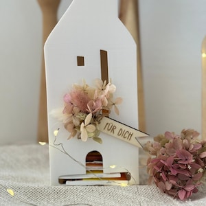 Könnte beinhalten: Ein weißer Kartenhalter in Hausform, verziert mit getrockneten Blumen in Rosa- und Beigetönen. Ein kleines Holzbanner mit dem Text "FÜR DICH" ist angebracht. Der Kartenhalter hat einen Schlitz zum Einstecken von Karten.