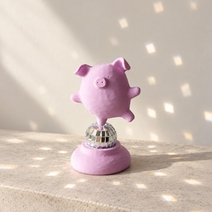 Könnte beinhalten: Eine hellviolette Schweinefigur mit matter Oberfläche, die auf einer kleinen, kugelförmigen, silbernen Discokugel balanciert. Die Discokugel ist auf einem runden, hellvioletten Sockel befestigt, der in Farbe und Textur der Schweinefigur entspricht.