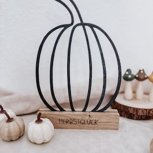 Może przedstawiać: Czarny kontur dyni na drewnianej podstawie z napisem "HERBSTGLÜCK". Dynia to element dekoracyjny, idealny do dekoracji w jesiennej tematyce. Małe dekoracyjne dynie na pierwszym planie.