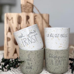 May include: Three speckled ceramic mugs with different sayings. The mugs are stacked on top of each other. The top mug says "La Vie Est Belle", the middle mug says "Not Your Ernst", and the bottom mug says "Et Voila Ta Realite".