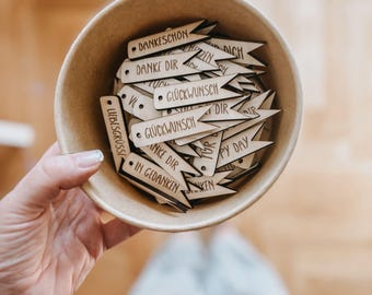May include: A beige bowl filled with small, wooden, arrow-shaped tags. The tags have various German phrases etched on them, such as "Danke Dir" and "Glückwunsch." The bowl is held in a person's hand, with a blurred background.