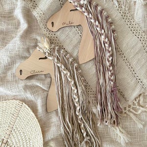 Könnte beinhalten: Zwei hölzerne Pferdekopf-Ornamente mit geflochtenem Garnhaar. Das Pferd links trägt den Namen "Clara" und das Pferd rechts den Namen "Mila" geschrieben.