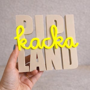 May include: A beige wooden sign with the words "PIPI KACKA LAND" in capital letters. The word "kacka" is in bright yellow cursive, while the other words are in a block font. The sign is held by a hand.