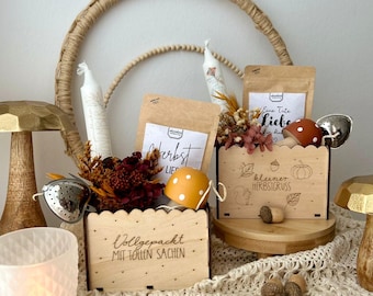 May include: A wooden box filled with autumn-themed items, including tea bags, a tea infuser, a candle, and decorative mushrooms. The box is labeled "Vollgepackt mit tollen Sachen" and the tea bags are labeled "Herbst Liebe" and "Kleiner Herbstgruss".