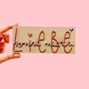 May include: A rectangular wooden sign with the words "sonst nichts" in red script lettering, with a small heart and a black accent. The sign is held against a pink background.
